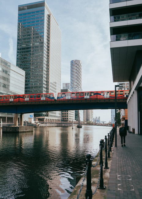 A modern urban scene featuring a river with calm, reflective water in the foreground, bordered by a paved walkway on the right side, which is lined with black metal posts and a chain-link barrier. To 
