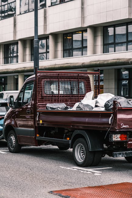 A small burgundy flatbed truck parked on an urban street in front of a modern office building with large glass windows and concrete facade. The truck's cargo area is filled with various types of waste