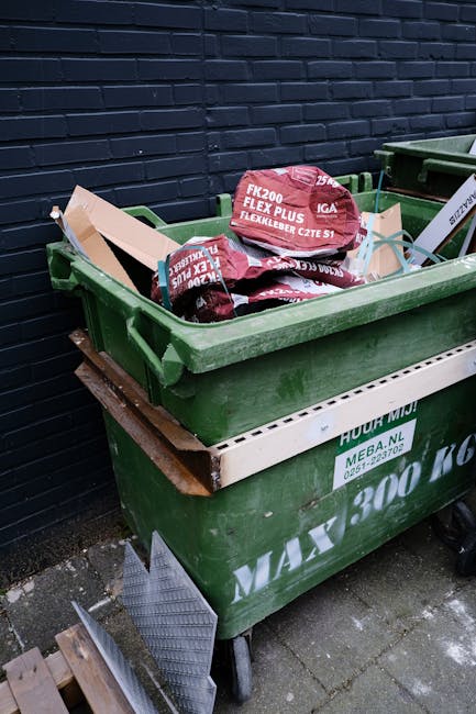 A small burgundy flatbed truck parked on an urban street in front of a modern office building with large glass windows and concrete facade. The truck's cargo area is filled with various types of waste
